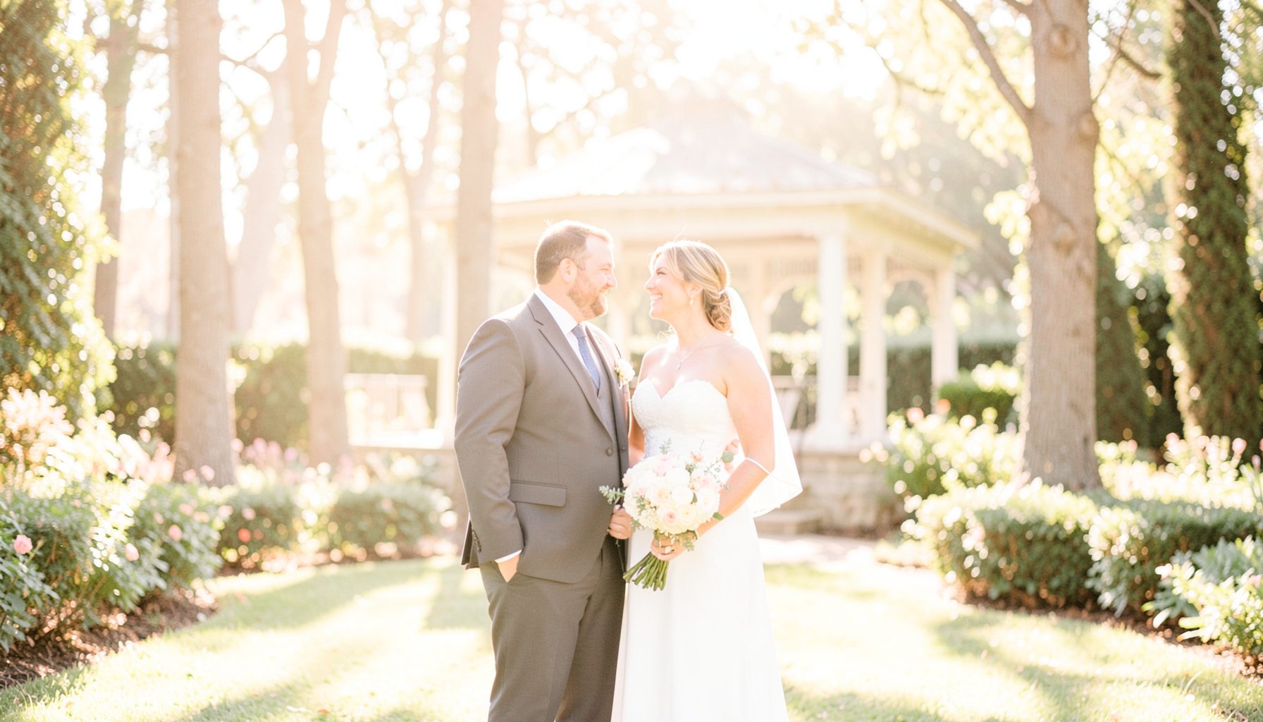 A bride and groom standing together in a sun-drenched garden, soft golden light filtering through tall trees and illuminating the gazebo behind them