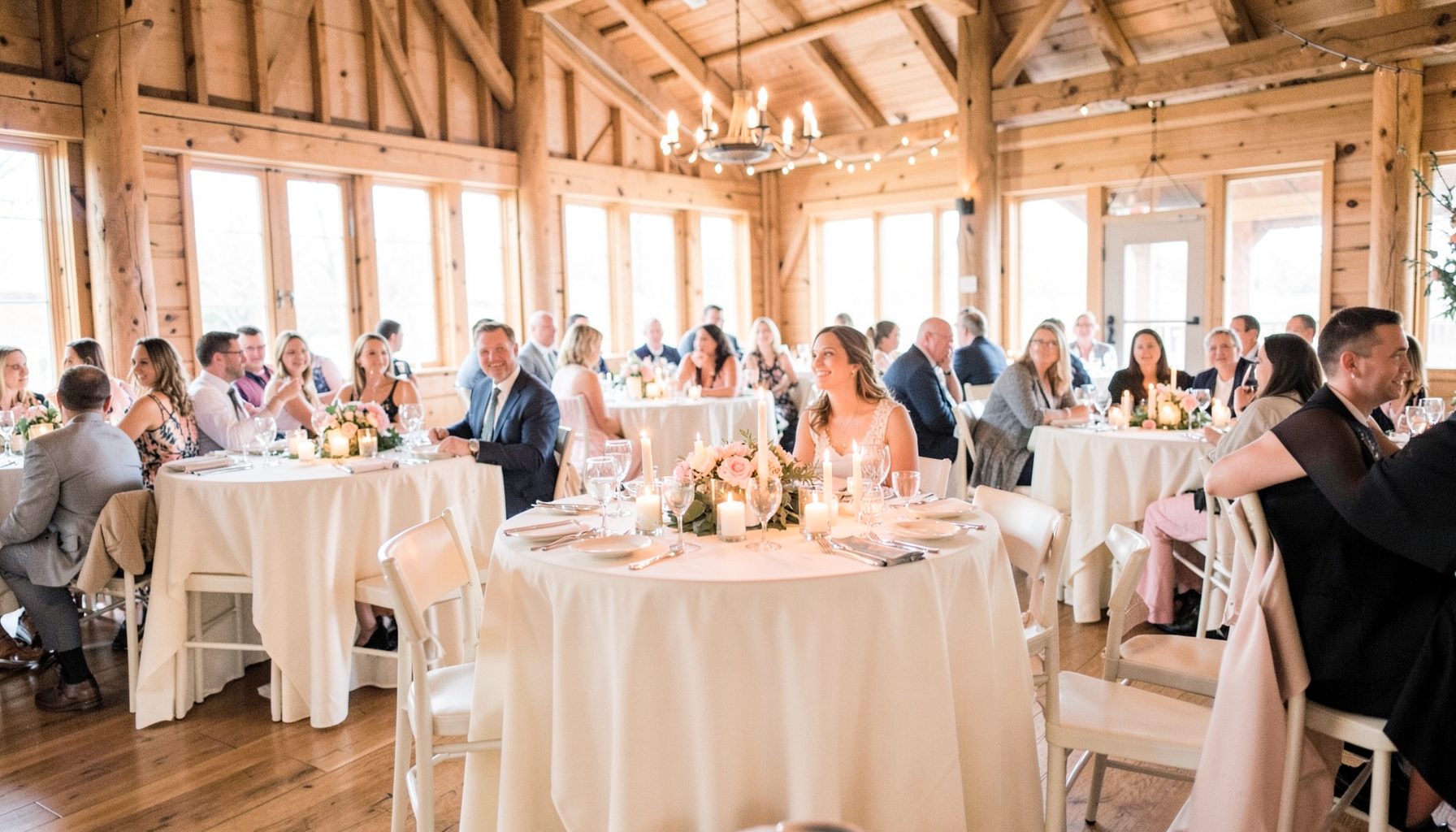 A warmly lit wedding reception inside a rustic lodge, with round tables draped in ivory linens, candlelight centerpieces, and guests enjoying the evening