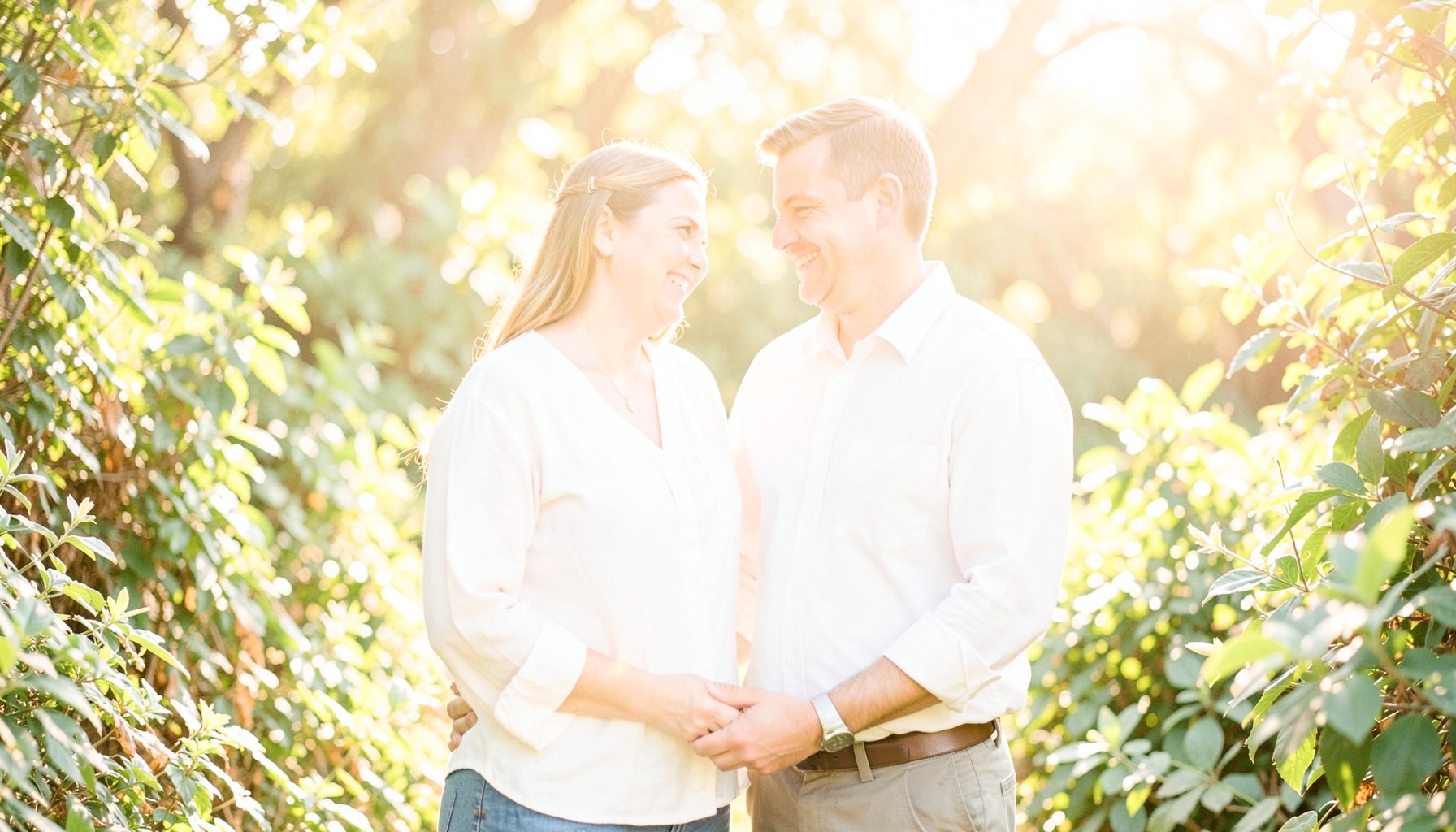 A bright, airy photograph of a couple standing together in soft natural light, surrounded by lush green foliage, with warm golden tones filtering through the trees