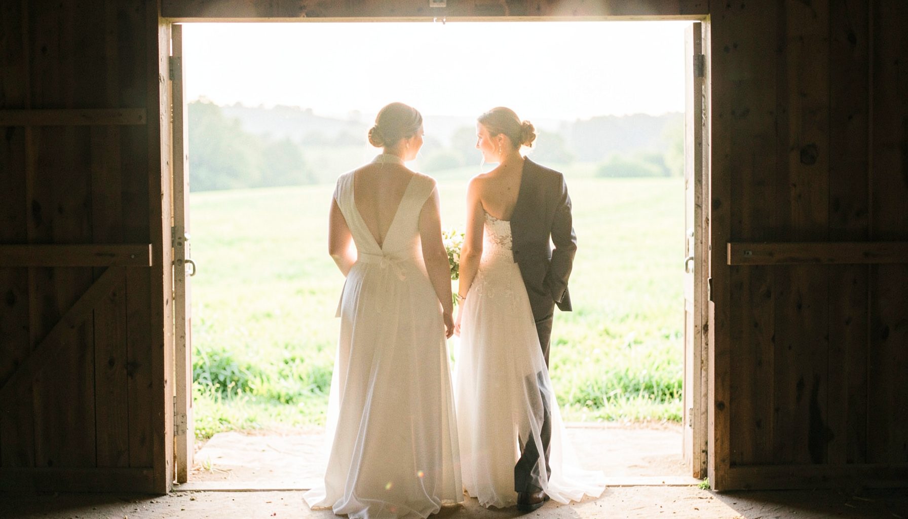 A bride and groom standing in a sun-drenched barn doorway, backlit by golden afternoon light, with rolling green fields beyond