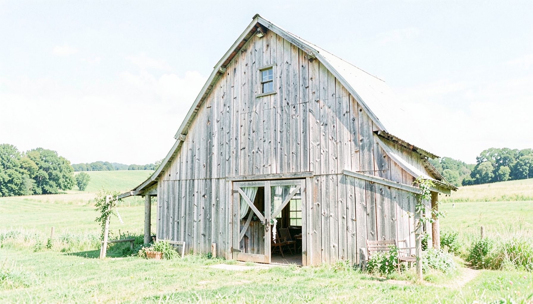 A sunlit rustic barn venue with weathered wood siding, surrounded by rolling green fields under a soft blue sky