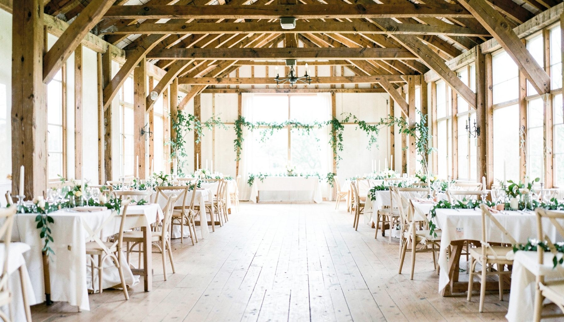 A sunlit rustic barn interior with exposed wooden beams, wide plank floors, and soft natural light streaming through tall windows